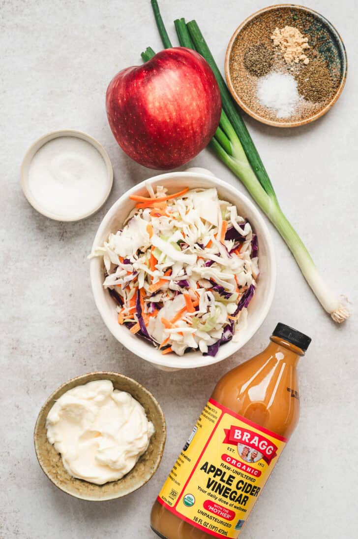 Ingredients on a light background, including shredded vegetables, apple cider vinegar, apple, green onion, mayonnaise and spices.