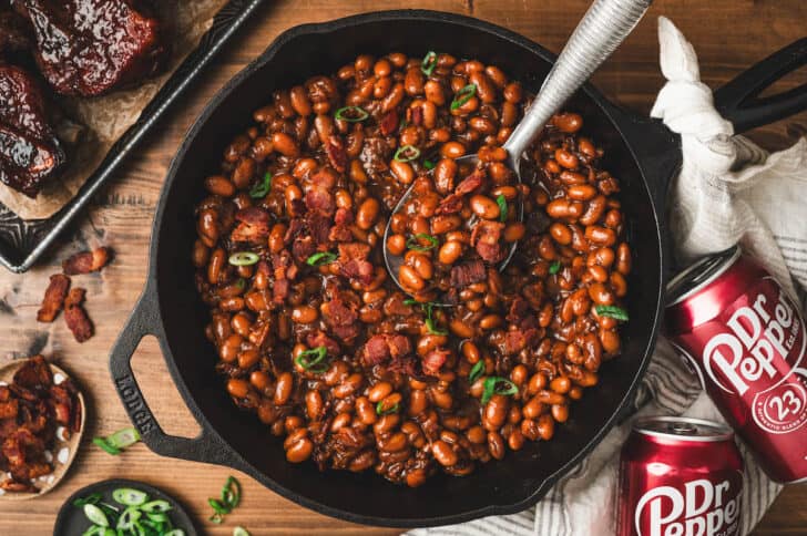 A cast iron skillet filled with baked beans with Dr. Pepper, with a spoon in them.