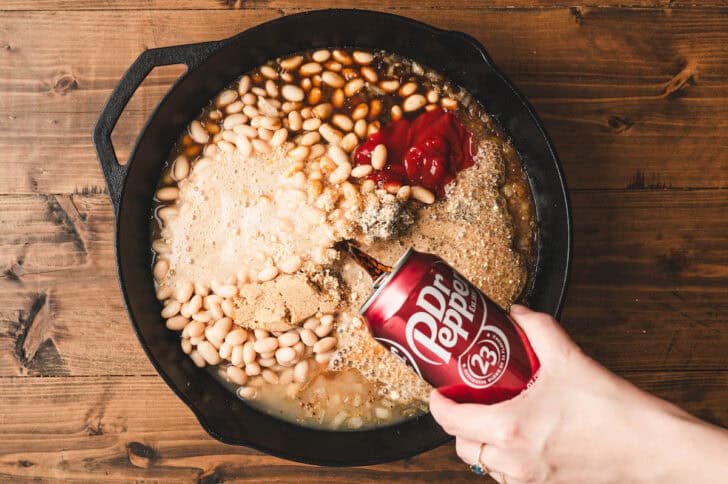 A hand pouring a can of Dr. Pepper into a cast iron skillet with beans and other ingredients.