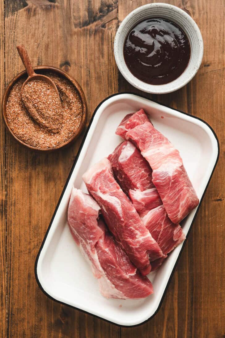 A plate of raw pork, with bowls of spices and sauce, on a wooden surface.