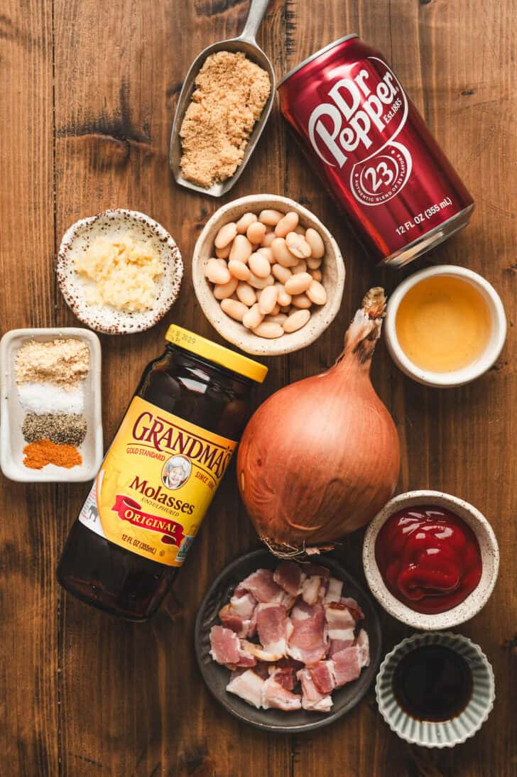 Ingredients on a wooden surface, including white beans, molasses, onion, bacon, Dr. Pepper and herbs and spices.