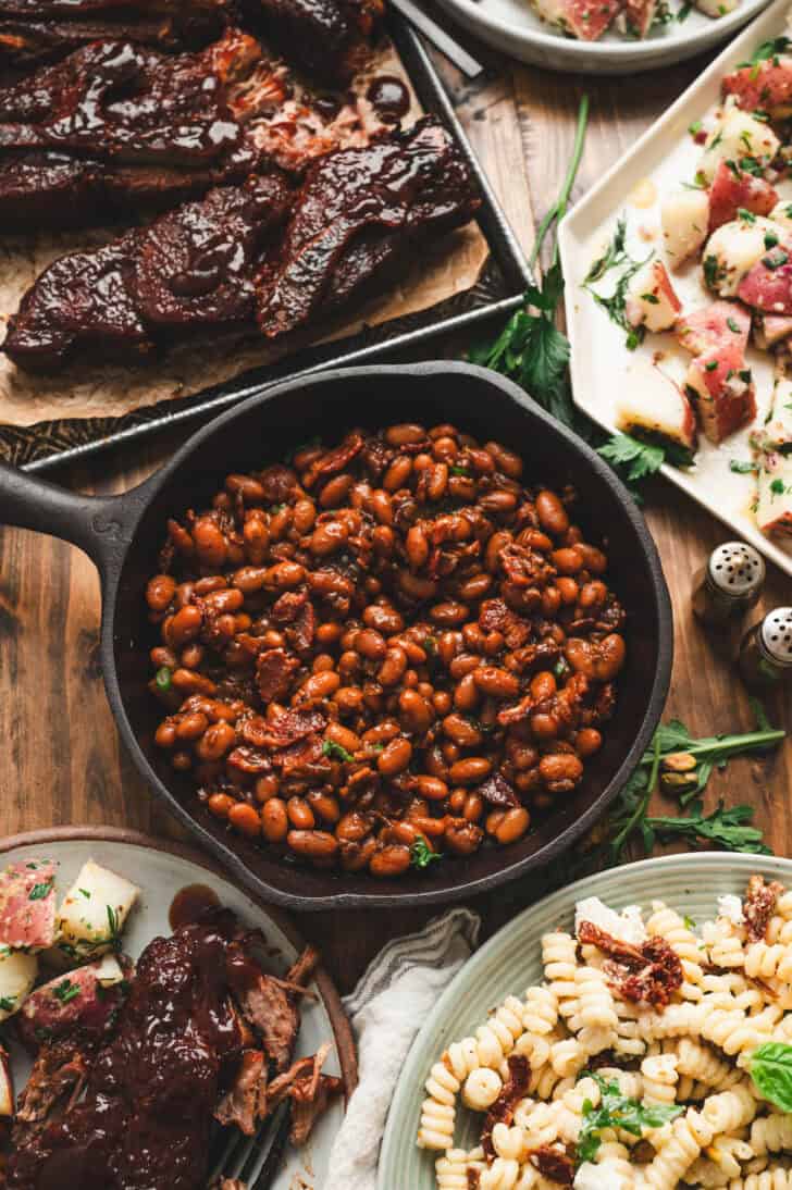 A skillet filled with a Dr. Pepper baked beans recipe, surrounded by ribs and other side dishes.