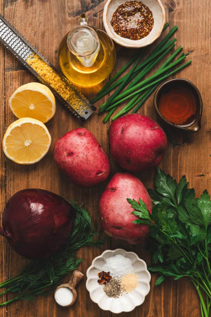 Ingredients on a wooden background, including red potatoes, lemons, onion, herbs, spices and oil.