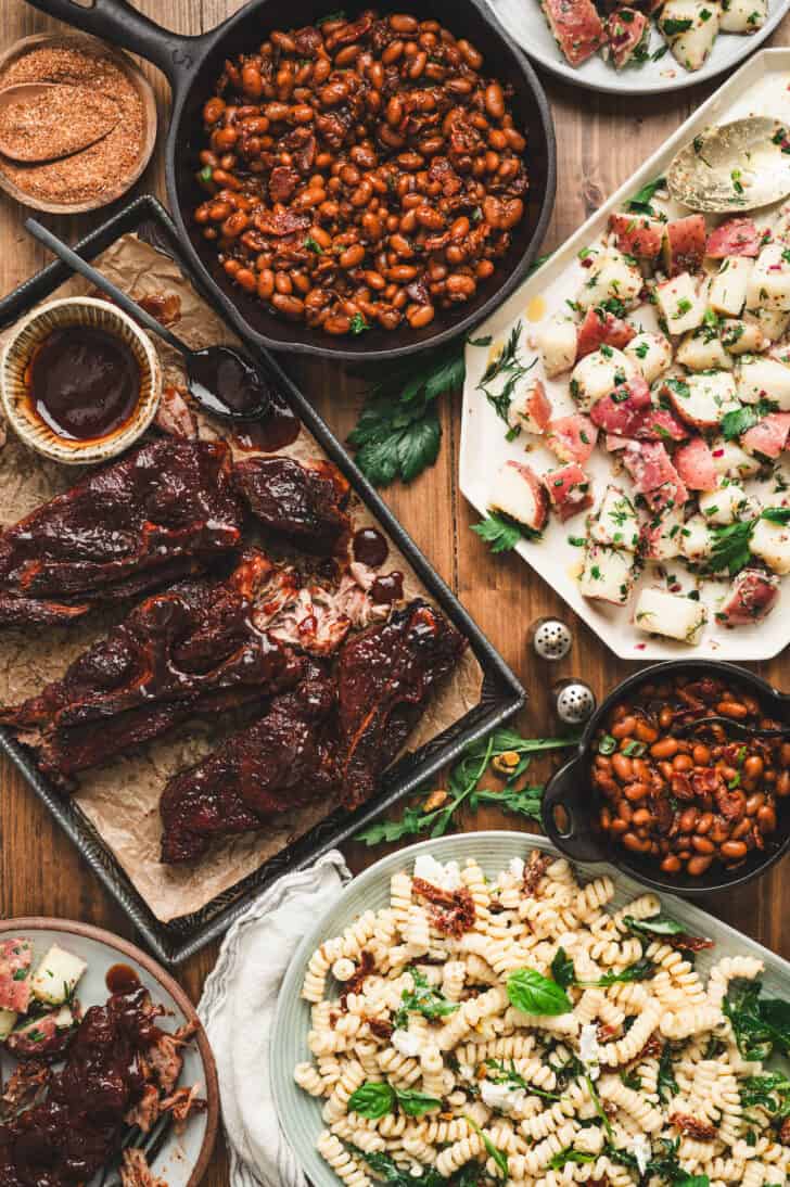 A dinner table filled with a country ribs recipe, baked beans, potato salad and pasta salad.