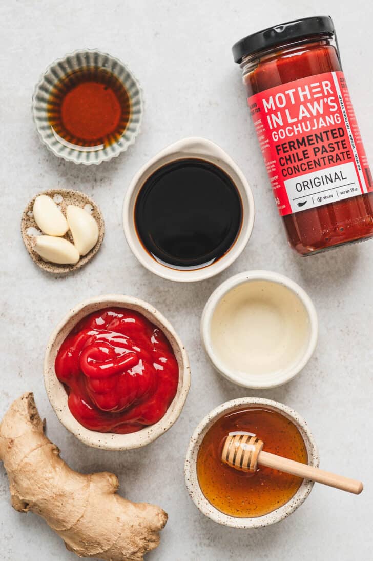 Ingredients on a light surface, including gochujang, honey, ketchup, garlic and ginger.
