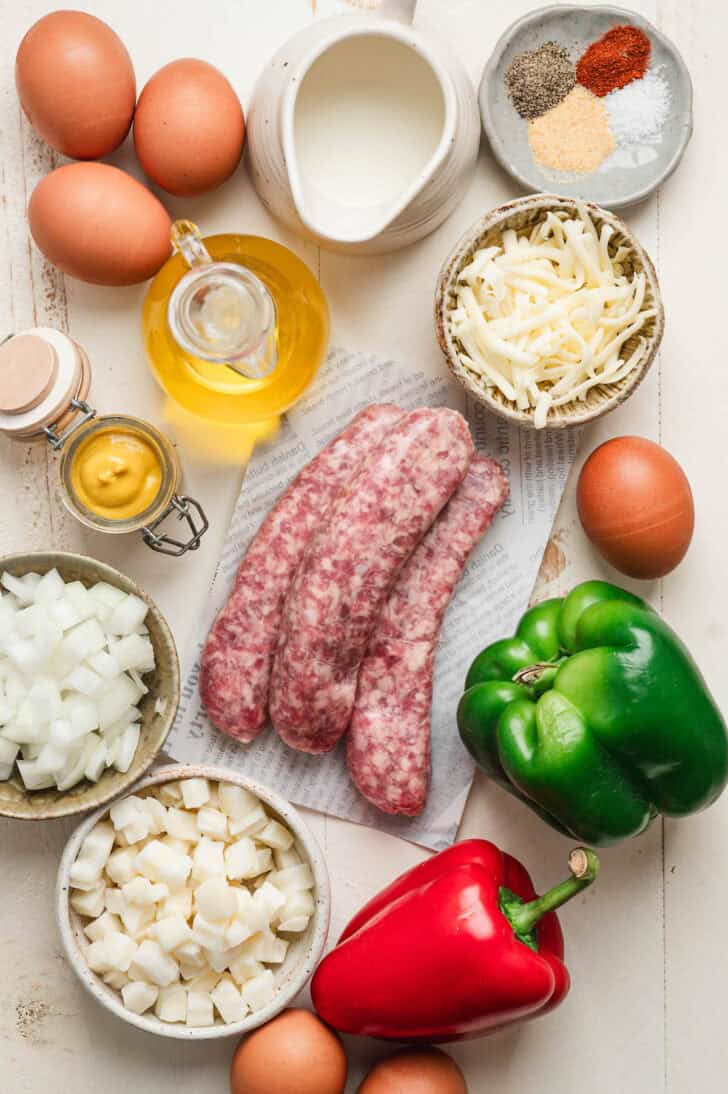 Ingredients on a light surface, including raw sausage links, bell peppers, eggs, cheese and spices.