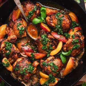 Overhead view of crispy skillet chicken thighs and peach wedges in a cast iron pan, topped with chopped green herb sauce and fresh basil leaves, with a spoon drizzling pan sauce over the top.