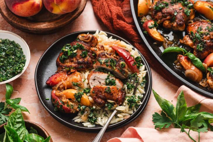 Sliced chicken thigh served over orzo pasta with peach wedges and herb sauce on a black plate, with a bowl of green herb sauce, fresh basil and parsley, and the skillet of chicken visible in the background.