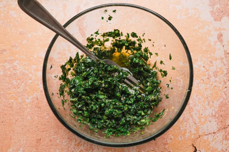 Close-up overhead view of a glass bowl containing finely chopped fresh herb sauce (gremolata-style) with olive oil, being mixed with a fork.