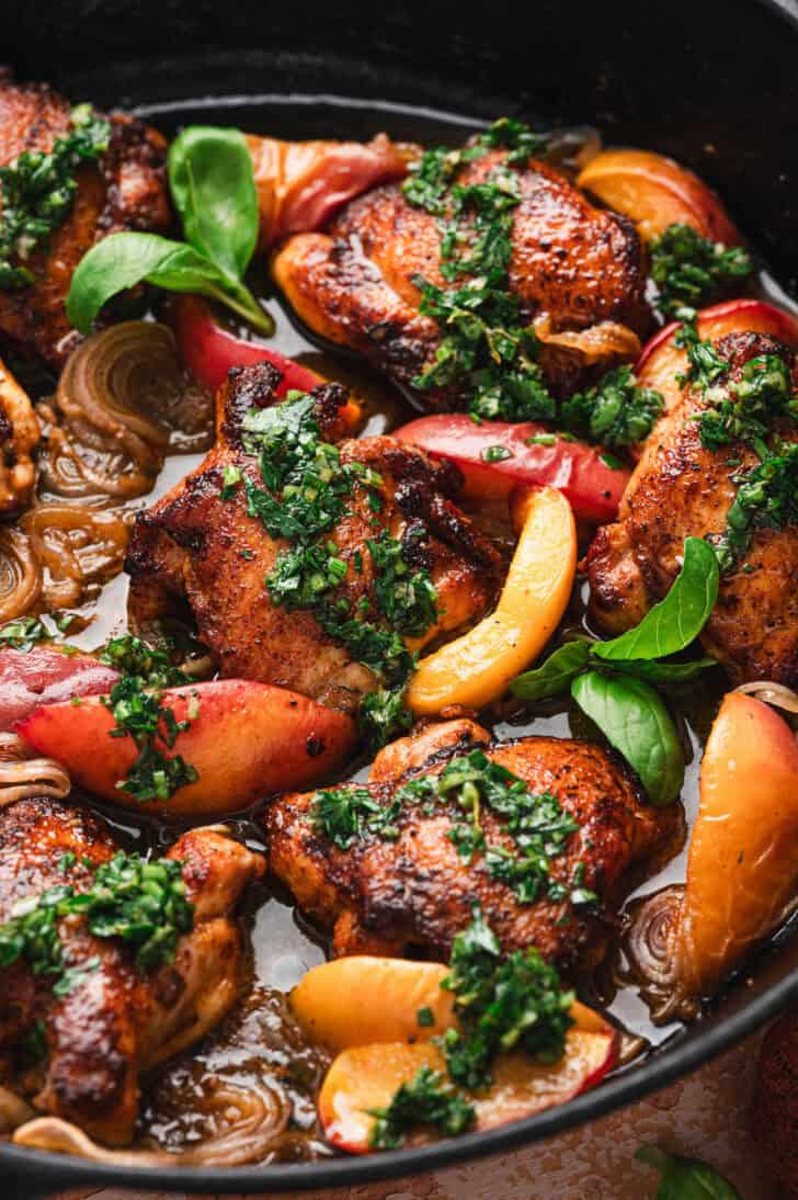 Close-up of golden-brown chicken thighs nestled among peach wedges and shallots in a cast iron skillet, generously topped with chopped herb sauce and garnished with fresh basil leaves.