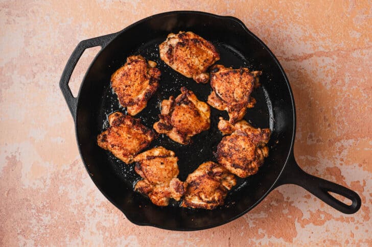 Overhead view of eight seasoned chicken thighs searing in a cast iron skillet, developing a golden-brown crust.