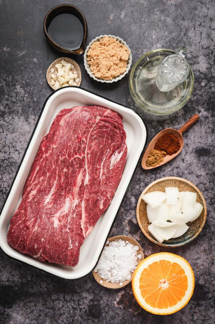 Ingredients on a textured gray surface, including raw beef, orange, onion, brown sugar and spices.