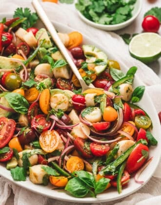 Heart of palm salad recipe with avocado, cherry tomatoes, red onion, basil, cilantro and jalapeno served on a platter with lime wedges.