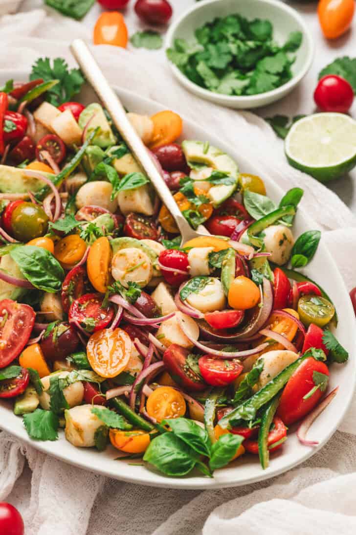 Heart of palm salad recipe with avocado, cherry tomatoes, red onion, basil, cilantro and jalapeno served on a platter with lime wedges.