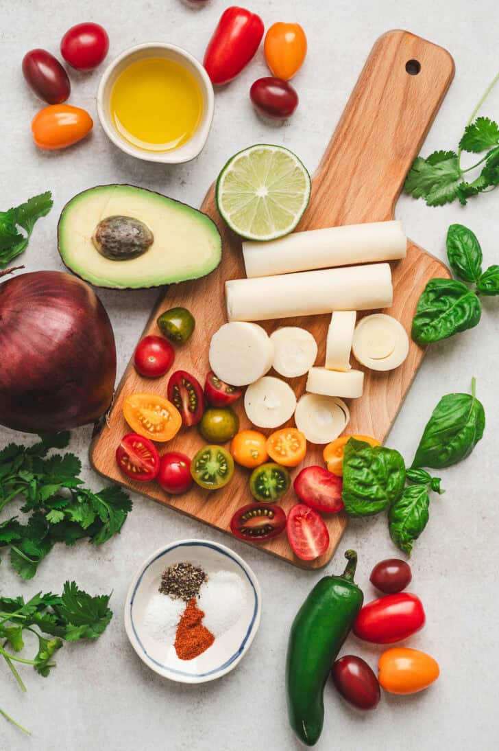 Ingredients for a hearts of palm salad recipe including hearts of palm, avocado, tomatoes, red onion, jalapeno, lime, herbs and olive oil.