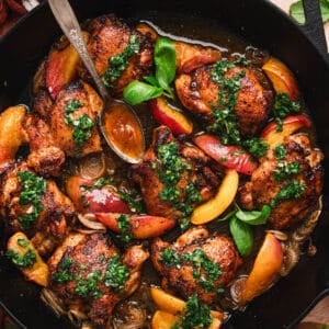 Vertical overhead shot of a cast iron skillet filled with glazed chicken thighs, peach slices, and shallots topped with herb sauce, with a spoon resting in the pan and whole peaches and a bowl of herb sauce visible at the edges.