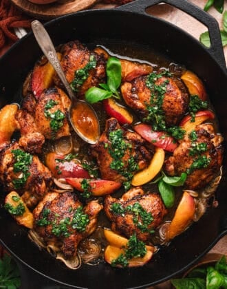 Vertical overhead shot of a cast iron skillet filled with glazed chicken thighs, peach slices, and shallots topped with herb sauce, with a spoon resting in the pan and whole peaches and a bowl of herb sauce visible at the edges.