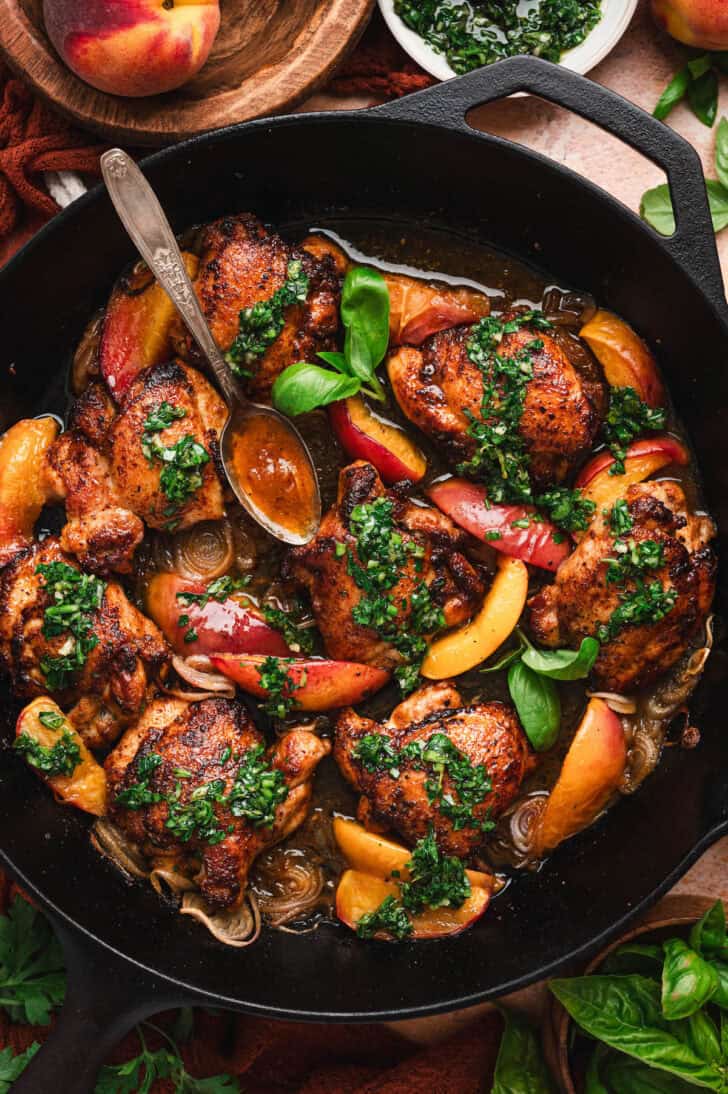 Vertical overhead shot of a cast iron skillet filled with glazed chicken thighs, peach slices, and shallots topped with herb sauce, with a spoon resting in the pan and whole peaches and a bowl of herb sauce visible at the edges.