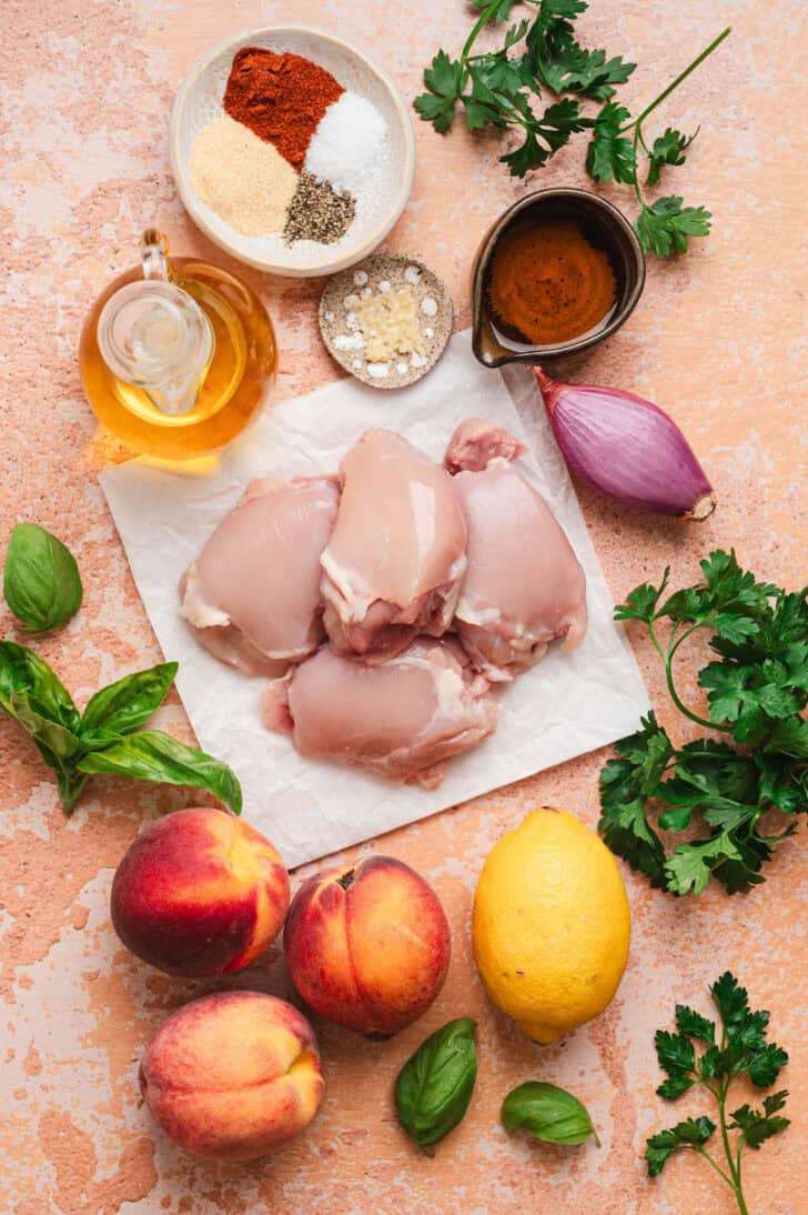 Flat-lay of raw ingredients for a chicken and peach dish, including boneless chicken thighs, whole peaches, a lemon, a shallot, garlic, a small bowl of spices (paprika, garlic powder, salt, pepper), honey and fresh basil and parsley.