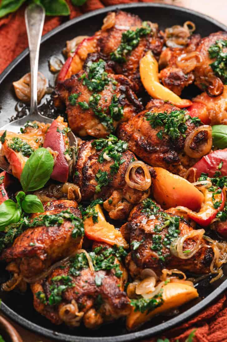 Close-up of glazed chicken thighs and peach wedges arranged on a dark plate, topped with chopped herb sauce and shallot rings, with a spoon and fresh basil leaves alongside.