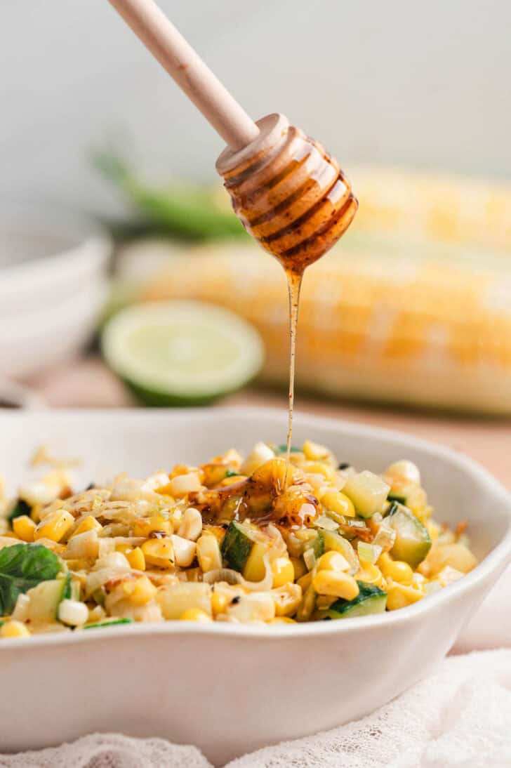 A honey dipper drizzling hot honey over a corn side dish recipe.