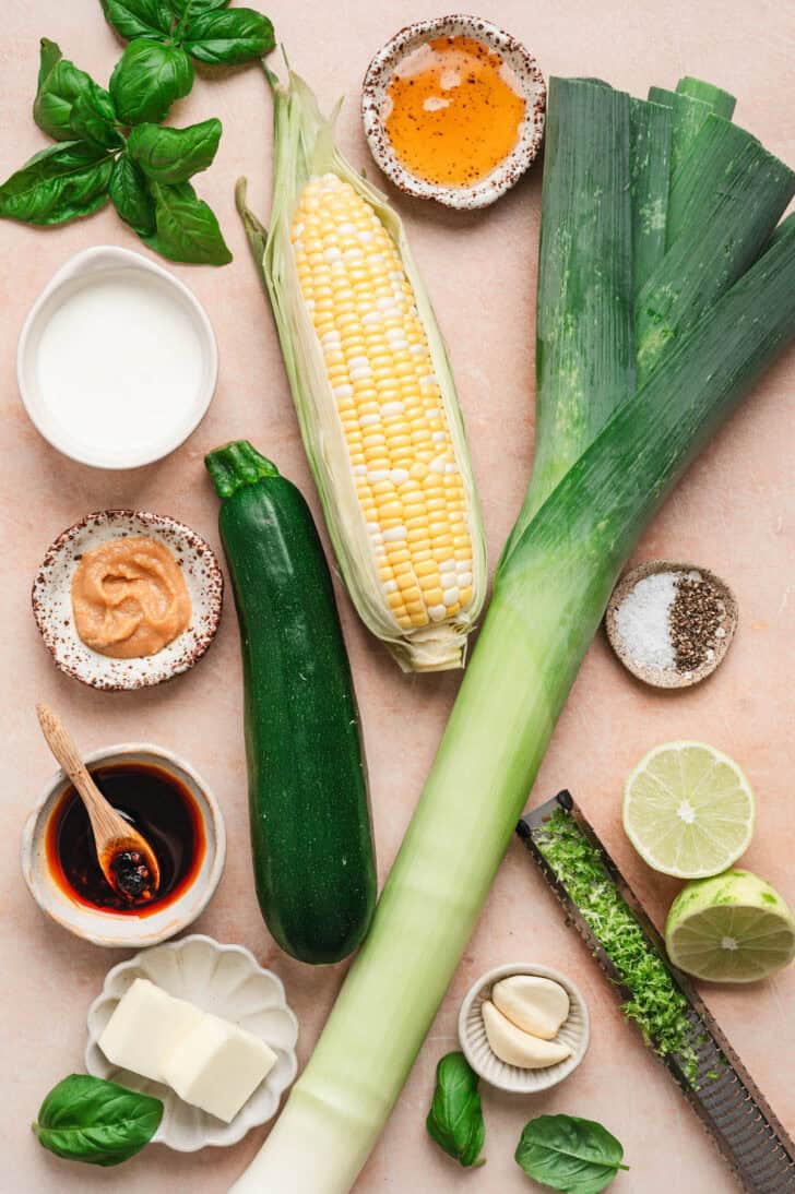 Ingredients on a light pink background, including corn, zucchini leeks, herbs and spices.