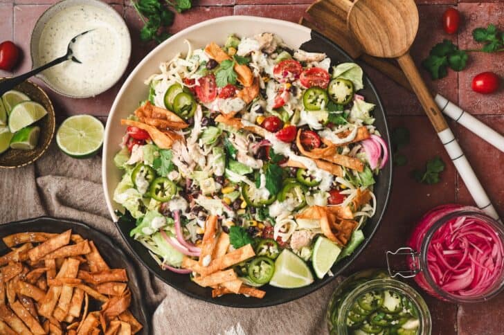 Overhead view of a black bean chicken salad topped with corn, shredded cheese, cherry tomatoes, pickled red onions, and crispy tortilla strips, drizzled with creamy ranch dressing and served with lime wedges and a jar of pickled jalapeños.