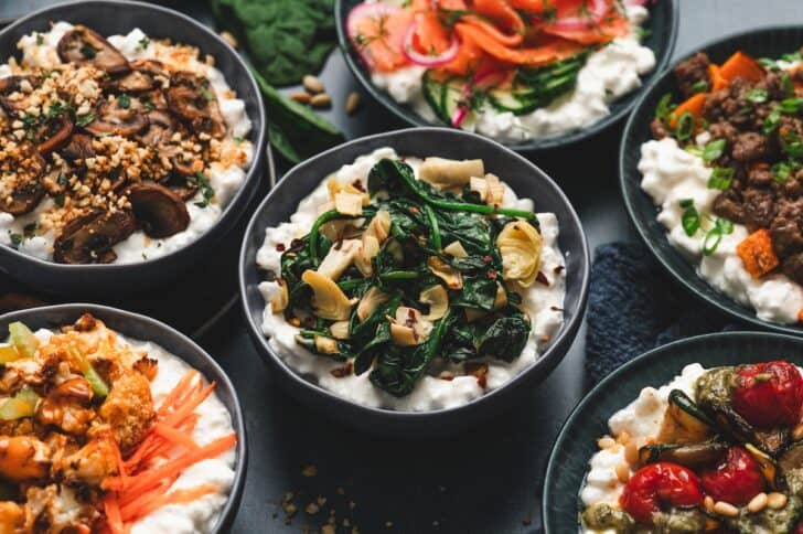 Close overhead view of a cottage cheese breakfast bowl spread, showing roasted mushroom, smoked salmon and dill, sausage and sweet potato, buffalo cauliflower, and pesto zucchini and tomato bowls arranged together.
