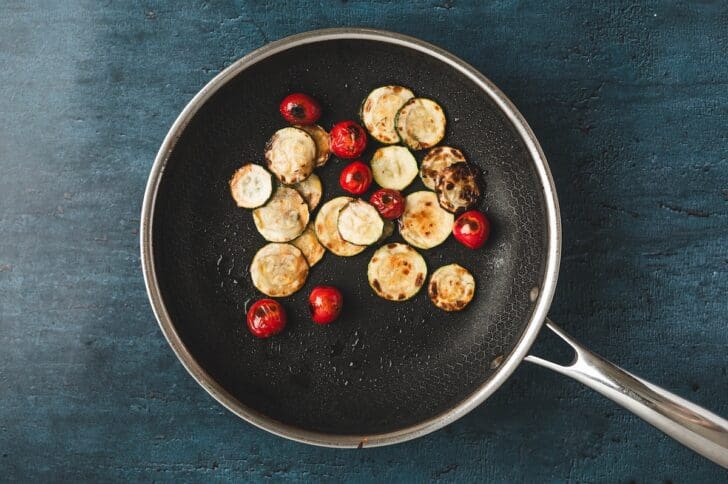 Overhead view of sliced zucchini and cherry tomatoes cooking in a skillet until softened and blistered.