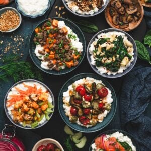 Overhead view of a cottage cheese breakfast spread featuring six bowls, including sausage and sweet potato, spinach artichoke, roasted mushroom, pesto zucchini and tomato, buffalo cauliflower, and smoked salmon and dill, arranged on a dark surface with fresh herbs and toppings.