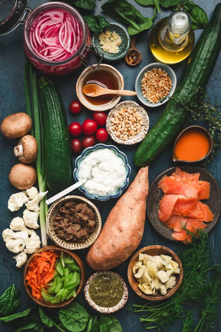 Flat-lay of ingredients for six savory cottage cheese bowls, including cottage cheese, smoked salmon, pickled red onions, sliced cucumbers, fresh dill, sweet potato, turkey sausage crumbles, mushrooms, panko breadcrumbs, fresh thyme, garlic, zucchini, cherry tomatoes, basil pesto, pine nuts, spinach, artichoke hearts, cauliflower, buffalo sauce, shredded carrots, and sliced celery.