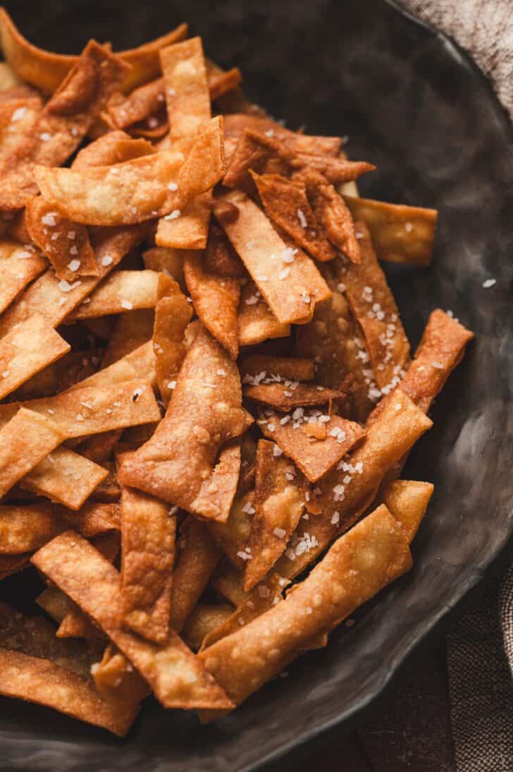 Close-up of crispy tortilla strips piled in a dark stone bowl and sprinkled with flaky sea salt.