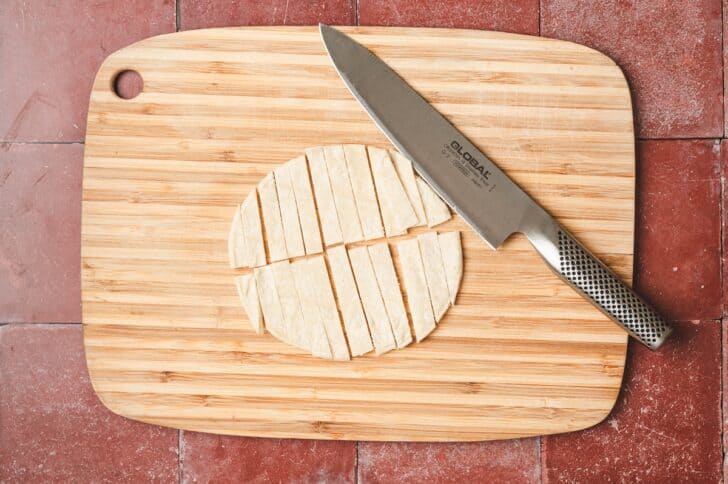 Overhead view of a corn tortilla sliced into thin strips on a wooden cutting board with a chef's knife.