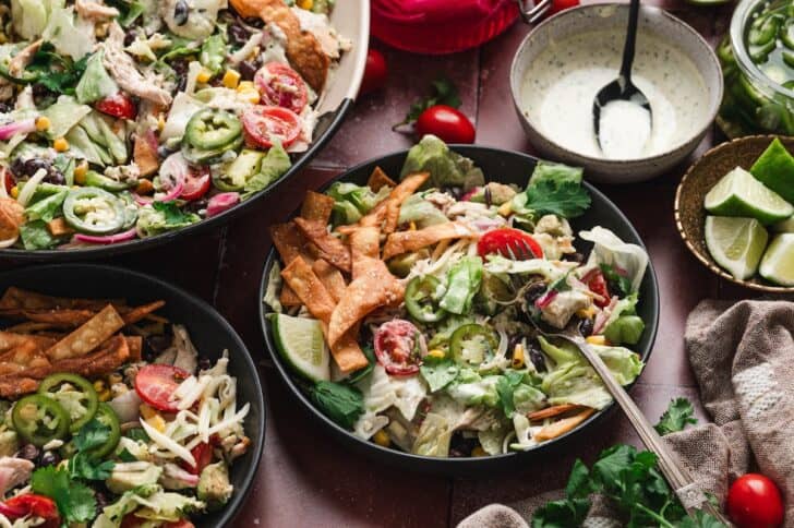 Overhead view of bowls of salad topped with homemade tortilla strips, black beans, jalapeño slices, and shredded chicken, served with a bowl of creamy dressing and lime wedges.