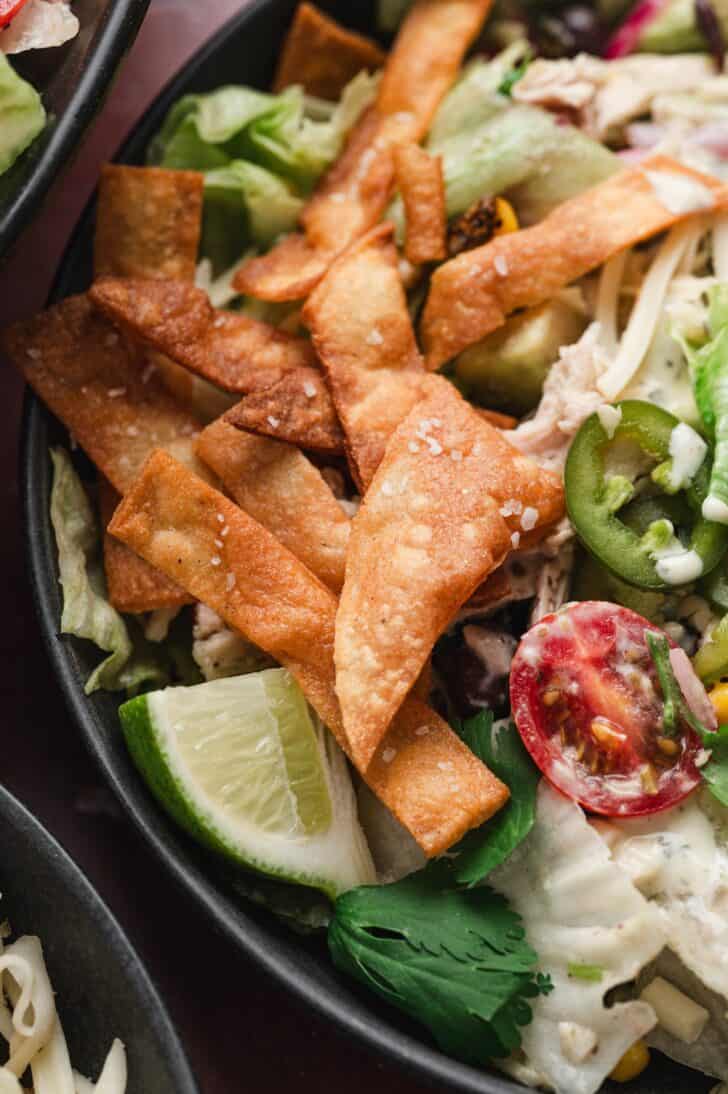 Close-up of a bowl from this tortilla strips recipe, showing crispy strips over salad greens with lime wedges, cherry tomatoes, jalapeño slices, and fresh cilantro.