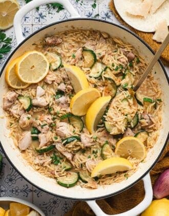 Overhead view of lemon chicken orzo in a white Dutch oven, with tender chicken, sautéed zucchini, and orzo pasta topped with lemon slices, parsley, and shaved parmesan.