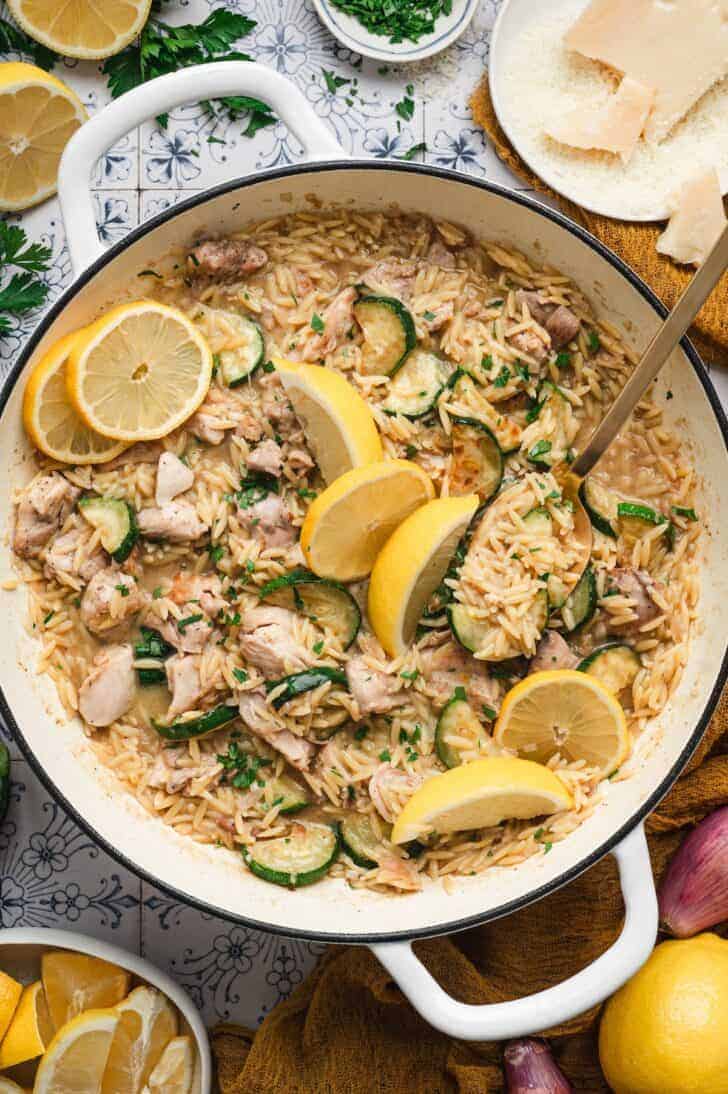 Overhead view of lemon chicken orzo in a white Dutch oven, with tender chicken, sautéed zucchini, and orzo pasta topped with lemon slices, parsley, and shaved parmesan.
