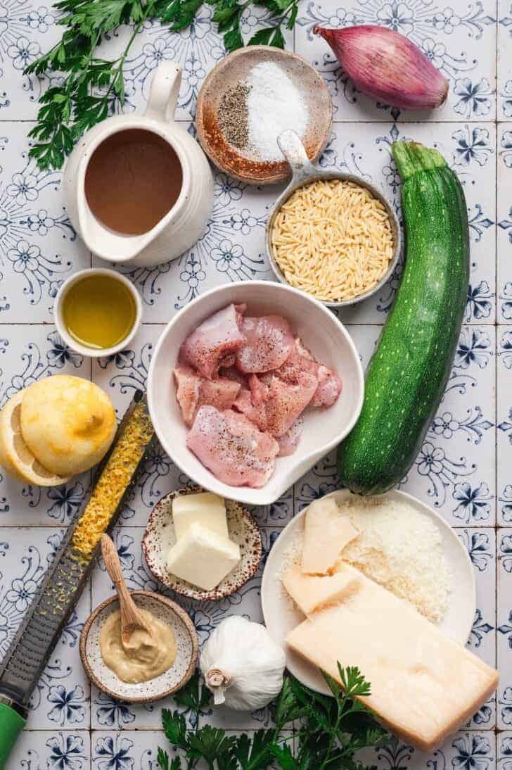 Flat-lay of ingredients for lemon chicken orzo, including raw chicken thighs, orzo pasta, a zucchini, a lemon, a shallot, garlic, chicken broth, butter, Dijon mustard, parmesan cheese, and fresh parsley.