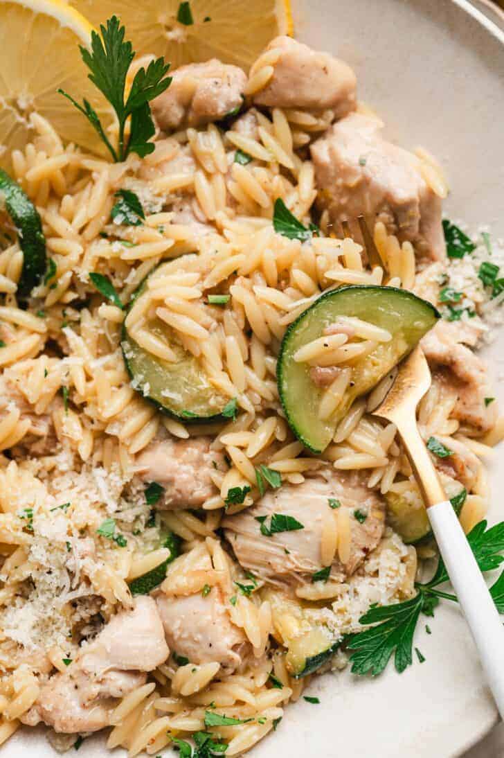 Close-up of a plate of lemon chicken orzo pasta with chicken pieces, sautéed zucchini, shaved parmesan, and fresh parsley, garnished with lemon slices.