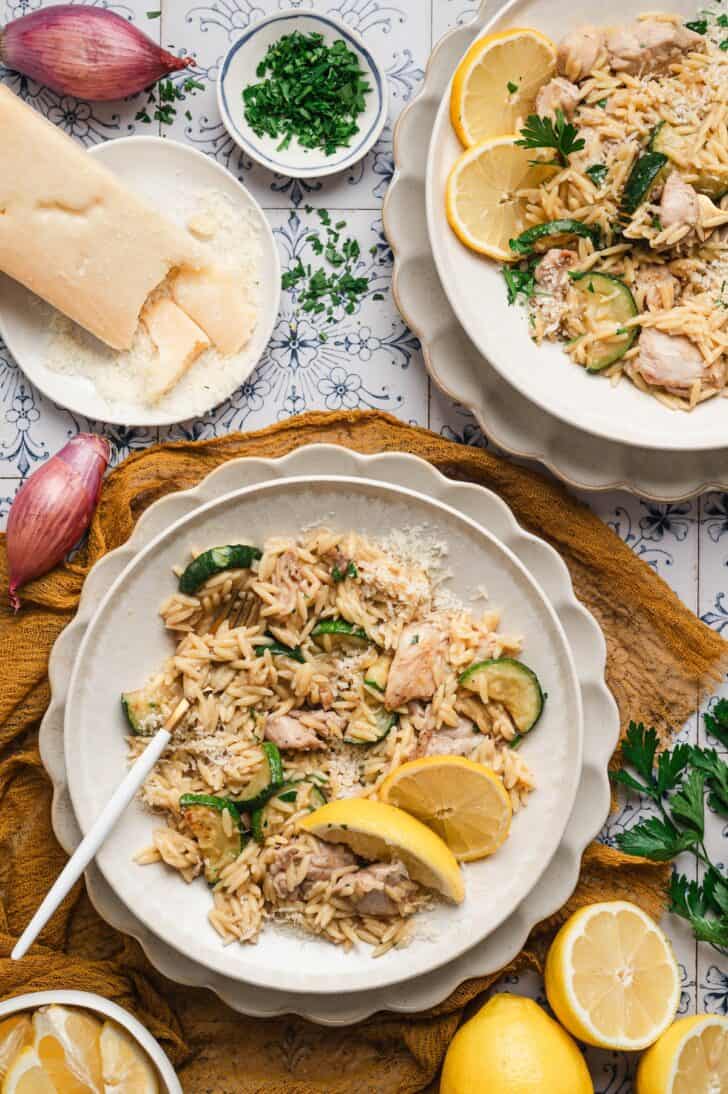 Overhead view of two plates of a lemon chicken orzo recipe topped with sautéed zucchini, shaved parmesan, chopped parsley, and lemon slices, with shallots and a block of parmesan nearby.