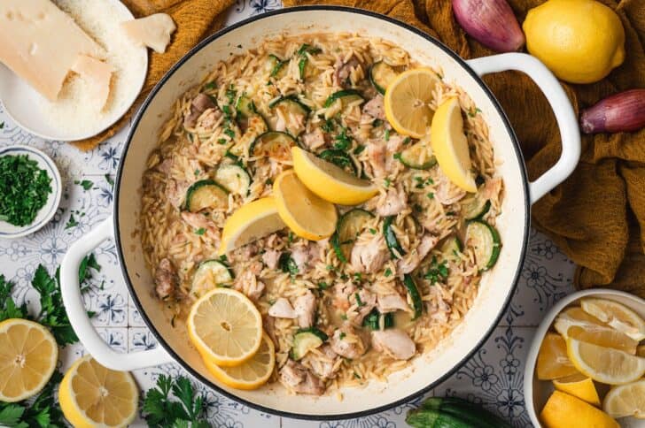 Overhead view of lemon orzo chicken in a white Dutch oven, garnished with lemon slices and fresh parsley, served with whole lemons and shallots alongside.