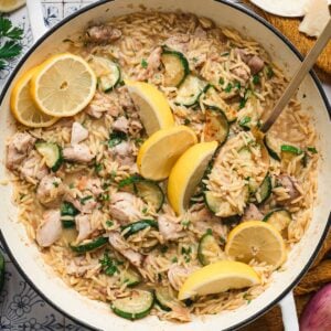 Overhead view of lemon chicken orzo in a white Dutch oven, with tender chicken, sautéed zucchini, and orzo pasta topped with lemon slices, parsley, and shaved parmesan.