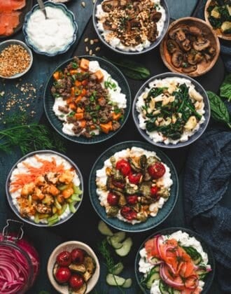 Overhead view of a savory cottage cheese breakfast spread with six bowls, including sausage and sweet potato, spinach artichoke, roasted mushroom, pesto zucchini and tomato, buffalo cauliflower, and smoked salmon and dill, surrounded by fresh herbs and ingredient garnishes.
