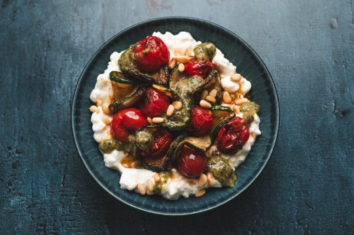 Overhead view of a pesto zucchini and tomato cottage cheese bowl, drizzled with basil pesto and topped with toasted pine nuts.