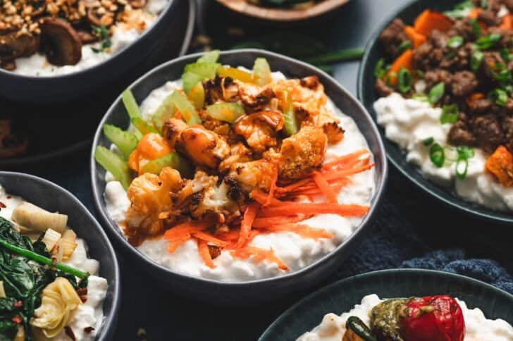 Close-up of a buffalo cauliflower cottage cheese bowl, topped with roasted cauliflower florets, buffalo sauce, shredded carrots, and sliced celery, with other cottage cheese bowls visible nearby.