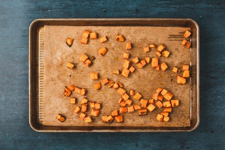 Overhead view of diced sweet potatoes roasted on a parchment-lined baking sheet until tender and caramelized.