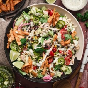 Overhead close-up of a southwest chicken salad recipe in a black bowl, layered with shredded chicken, black beans, corn, tomatoes, jalapeño slices, pickled red onion, shredded cheese, crispy tortilla strips, and cilantro, drizzled with ranch dressing and served with lime wedges.
