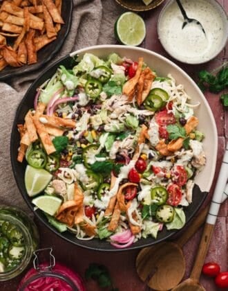 Vertical overhead shot of a southwest salad recipe in a large bowl, topped with shredded chicken, black beans, roasted corn, tomatoes, jalapeño slices, pickled onions, cheese, and crispy tortilla strips, served alongside tortilla chips, lime wedges, a jar of pickled onions, and a bowl of ranch dressing.