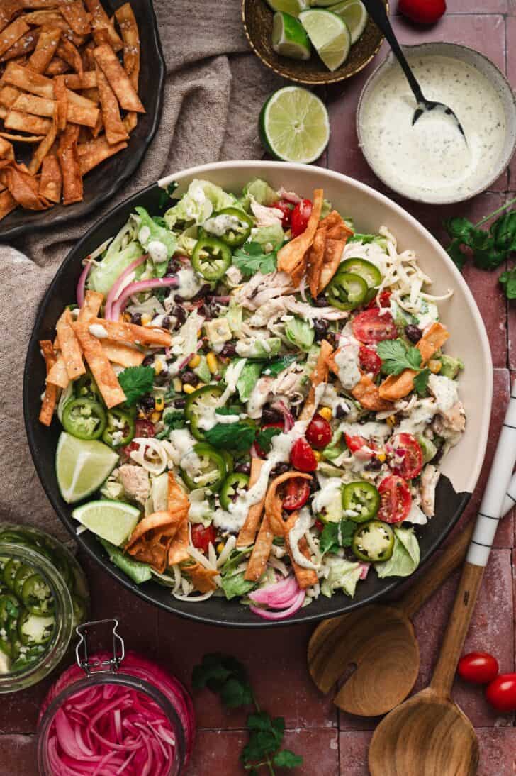 Vertical overhead shot of a southwest salad recipe in a large bowl, topped with shredded chicken, black beans, roasted corn, tomatoes, jalapeño slices, pickled onions, cheese, and crispy tortilla strips, served alongside tortilla chips, lime wedges, a jar of pickled onions, and a bowl of ranch dressing.