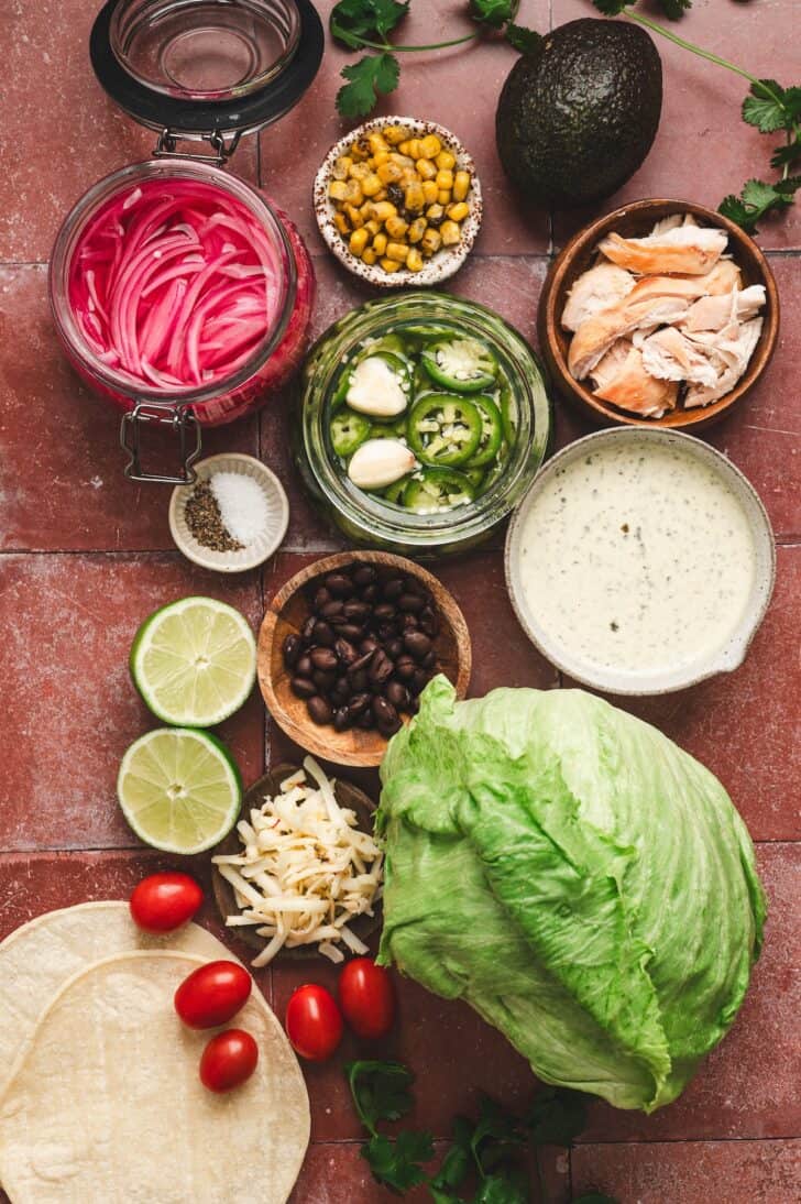 Flat-lay of ingredients for a southwest salad recipe, including pickled red onions, pickled jalapeños with garlic, roasted corn, shredded chicken, black beans, shredded cheese, halved limes, cherry tomatoes, corn tortillas, iceberg lettuce, and a bowl of creamy dressing.